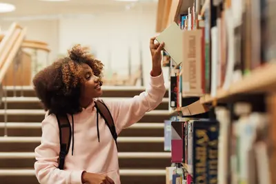 A smiling student with a backpack reaches up to pull a book from a library shelf.