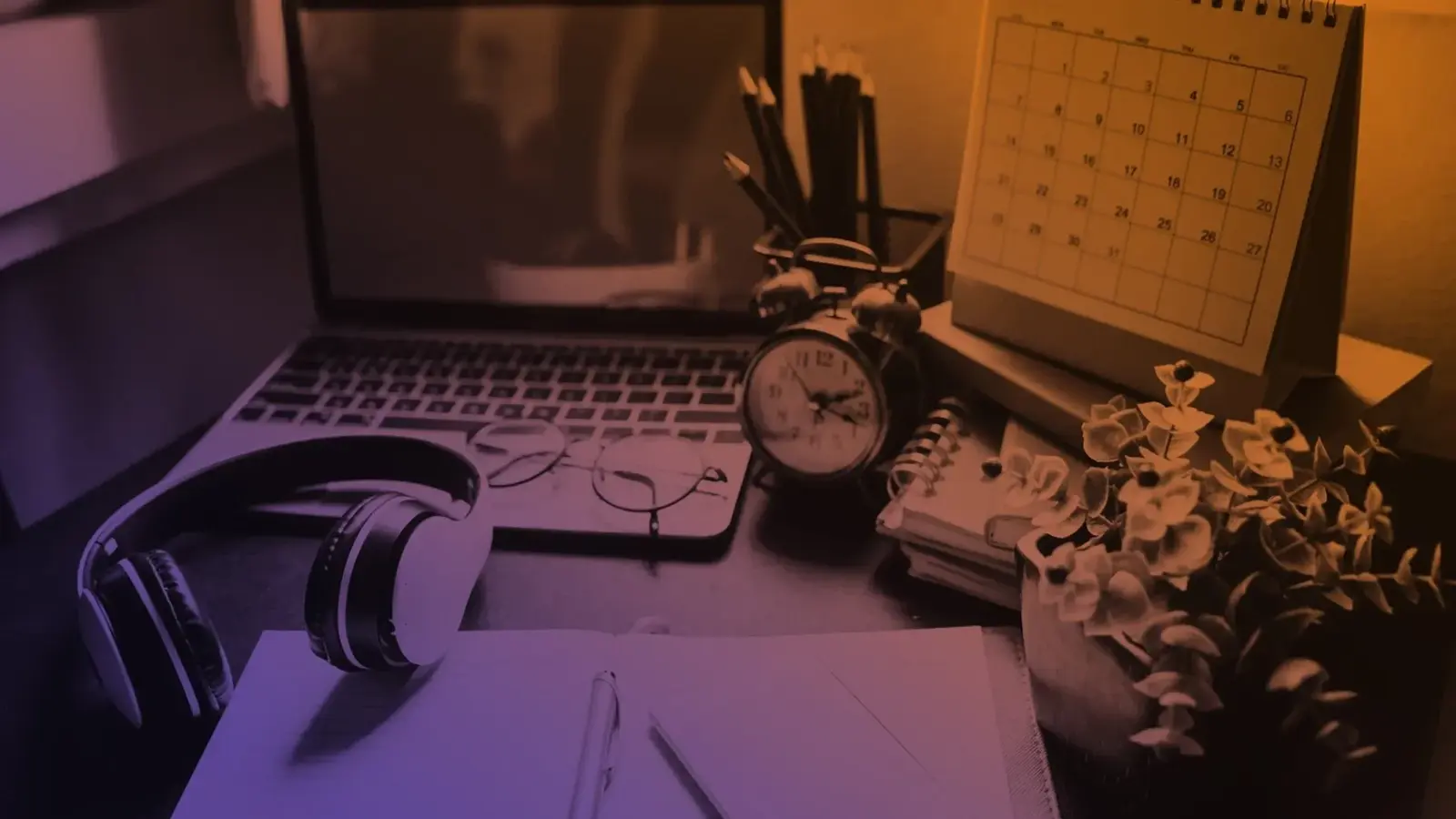 A desk with a laptop, headphones, glasses, alarm clock, notebooks, pencils, a desk calendar, and a plant