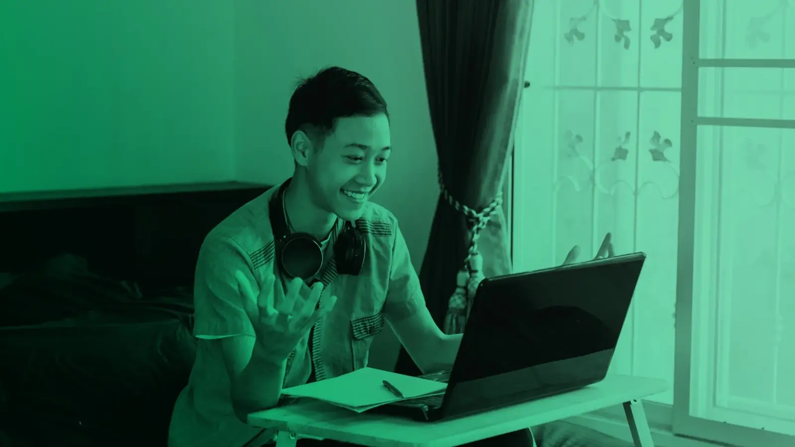 A smiling student with headphones around his neck gestures while looking at a laptop at a desk with an open notebook.