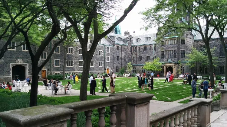 Students and guests gathered on a green quad outside a Gothic stone college building, seen from a stone balustrade.