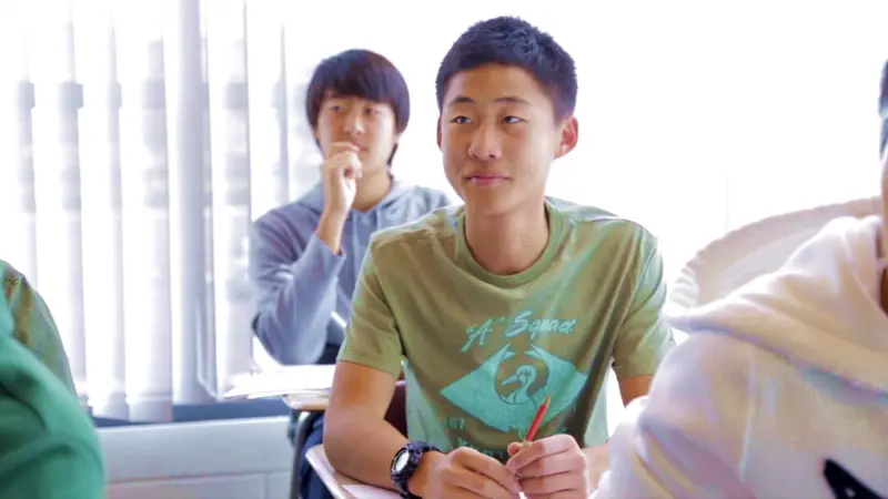 A student in a green T-shirt holding a pencil listens attentively in a bright classroom with classmates nearby.