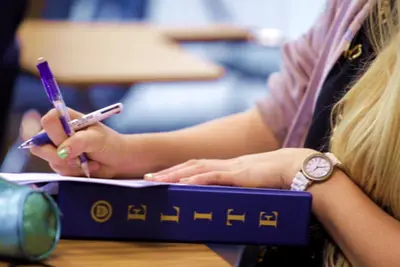 Student writing with a purple pen beside a blue binder labeled "ELITE" on a classroom desk