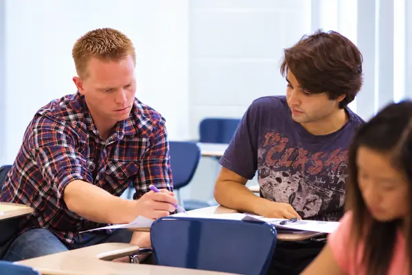 A tutor in a plaid shirt writes notes while a student watches at a classroom desk