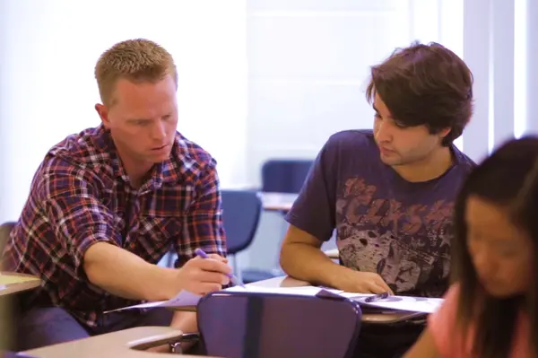 A tutor in a plaid shirt writes notes while a student watches at a classroom desk