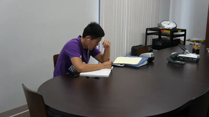 A student in a purple shirt with earbuds writes in a notebook at a large conference table.