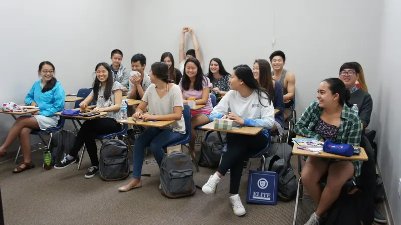Students sitting at desks in a classroom, smiling and laughing together, with backpacks on the floor.