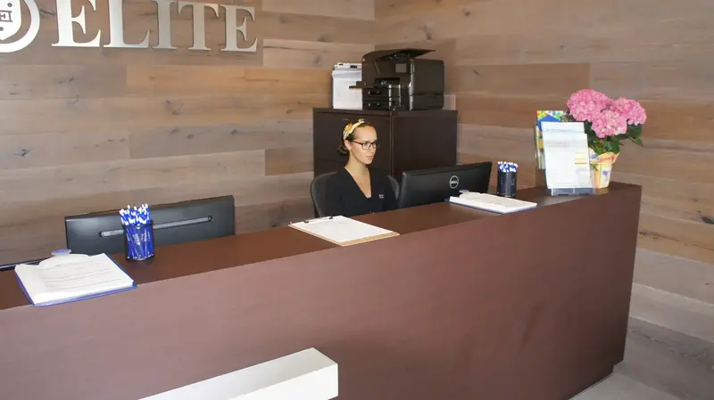 A receptionist sits behind a brown front desk in a wood-paneled lobby beneath an "ELITE" sign.