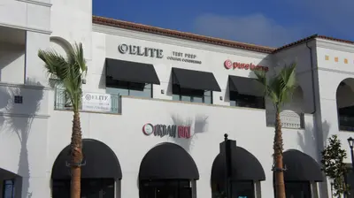 White Spanish-style shopping plaza with black awnings and palm trees, signs reading Elite Test Prep College Consulting, pure barre, and Urban Girl.