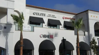 White Spanish-style shopping plaza with black awnings and palm trees, signs reading Elite Test Prep College Consulting, pure barre, and Urban Girl.