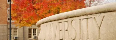 Stone campus wall carved with the word "UNIVERSITY" in front of a tree with red autumn leaves and a brick building