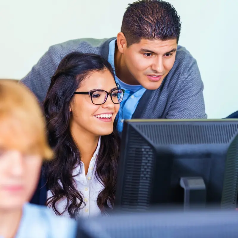 A smiling student in glasses looks at a computer monitor while a teacher leans in beside her