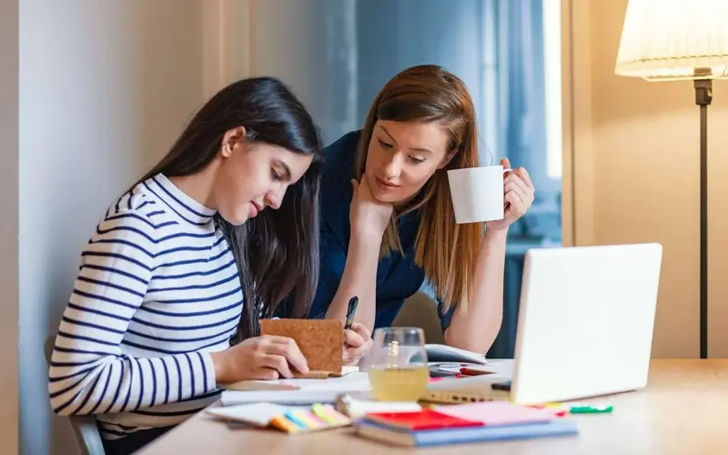 A tutor holding a mug leans over a desk to help a student taking notes beside a laptop and study materials.