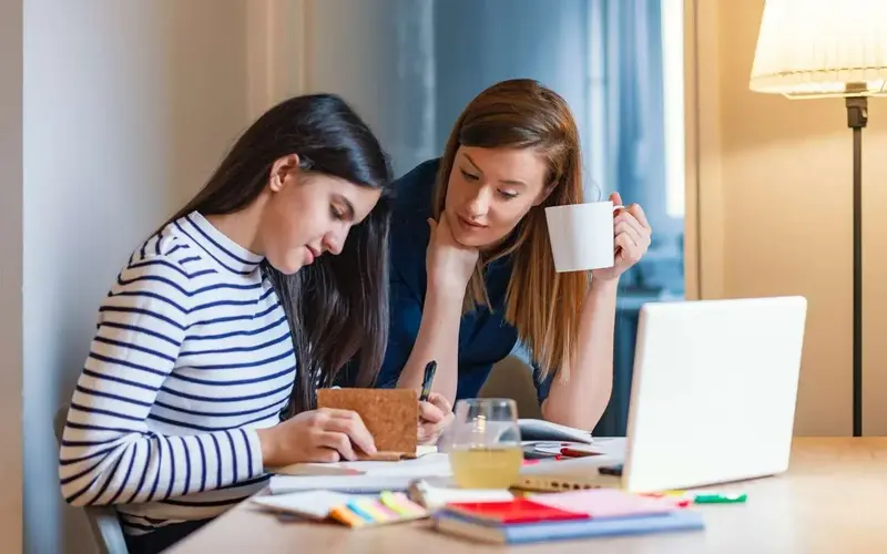 A tutor holding a mug leans over a desk to help a student taking notes beside a laptop and study materials.