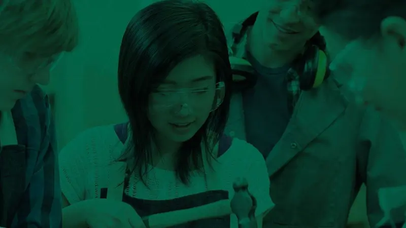 A student in safety goggles smiles while working on a project, with classmates gathered around her