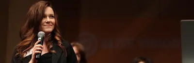 A smiling woman holding a microphone speaks on stage in front of an "Elite Vision" backdrop.
