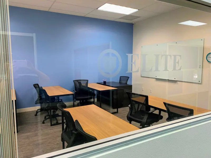 Classroom with wooden desks, black chairs, a whiteboard and blue wall, seen through a glass window with "Elite" logo