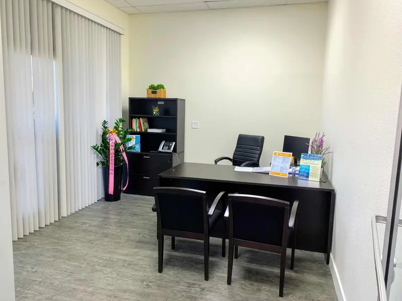 Office consultation room with a dark wood desk, chairs, bookshelf, plants, and vertical blinds along one wall.