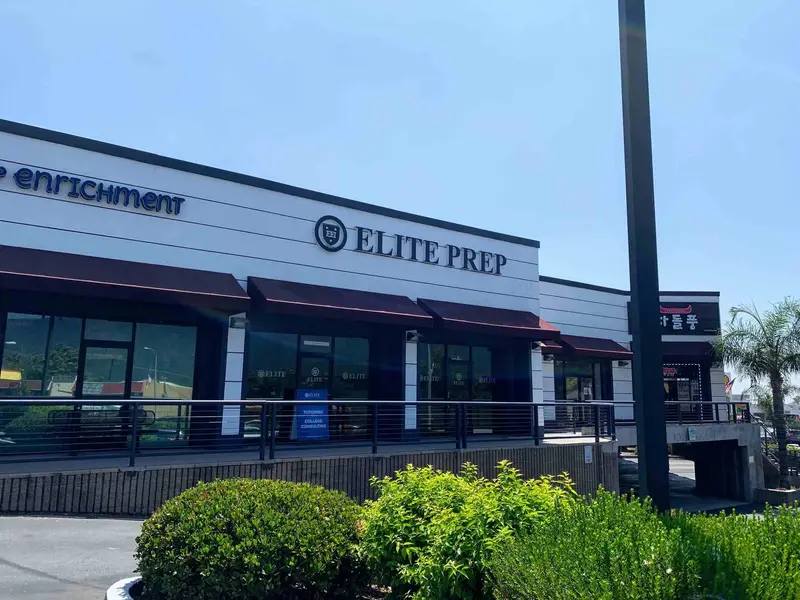 Storefront of a strip mall building with an "ELITE PREP" sign and dark awnings above windows, with shrubs in front.