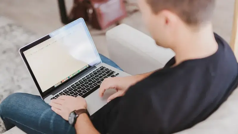 A young man sitting on a couch types on a laptop resting on his knees, with a blank document open.