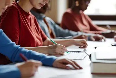 Students seated in a row at a long desk writing with pens on papers during a test