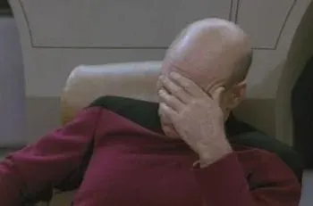 A bald man in a red uniform sits in a chair covering his face with his hand in a facepalm.