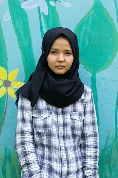 A young woman in a black hijab and plaid shirt stands before a teal wall painted with flowers and leaves.