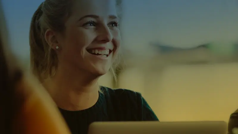 A smiling young woman with blonde hair sits in front of an open laptop screen