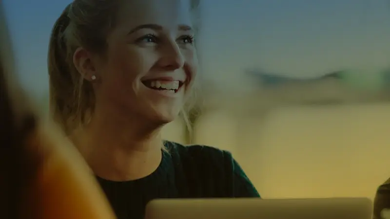 A smiling young woman with blonde hair sits in front of an open laptop screen