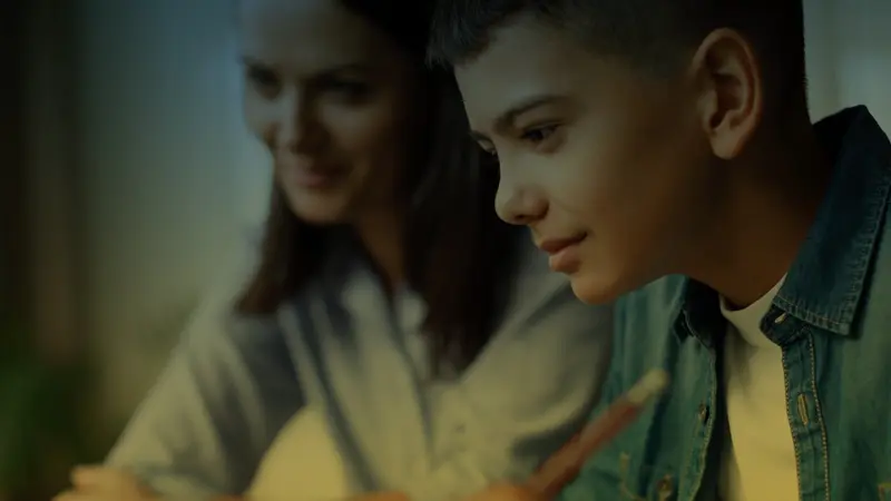 A boy in a denim jacket looks at a glowing screen while a woman smiles beside him