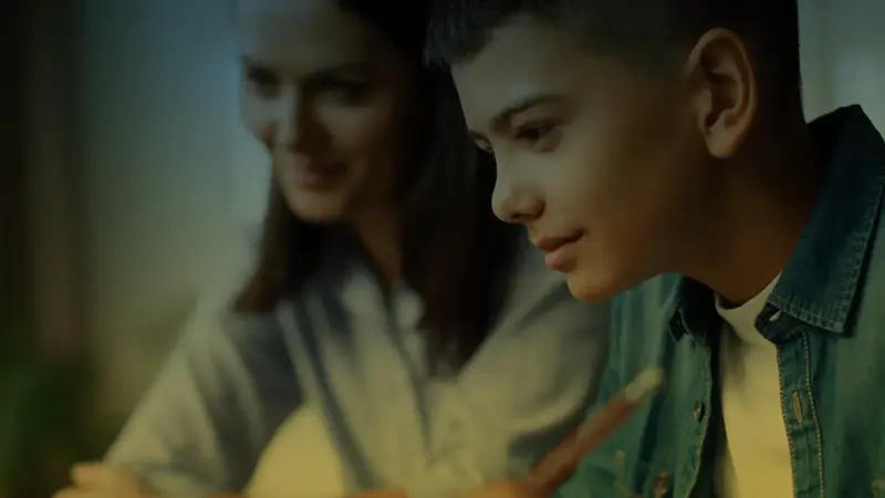 A boy in a denim jacket looks at a glowing screen while a woman smiles beside him