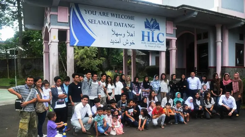 Large group of adults and children posing outside a building under a banner reading "You Are Welcome / Selamat Datang" with the HLC Hope Learning Center logo.