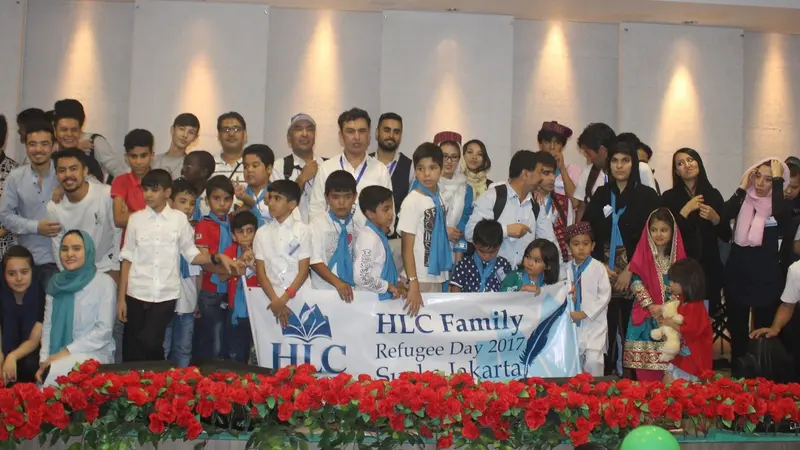 Large group of children and adults posing on stage behind a banner reading "HLC Family Refugee Day 2017", with red flowers in front.