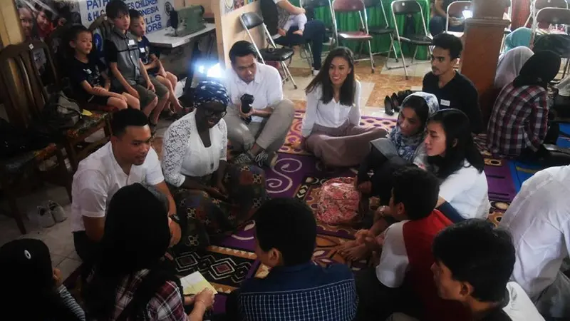Adults and children sit together in a circle on patterned rugs in a crowded room during a community gathering.