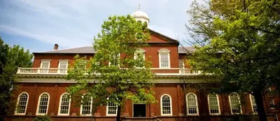 A red brick college building with a white cupola and arched windows, framed by leafy green trees.