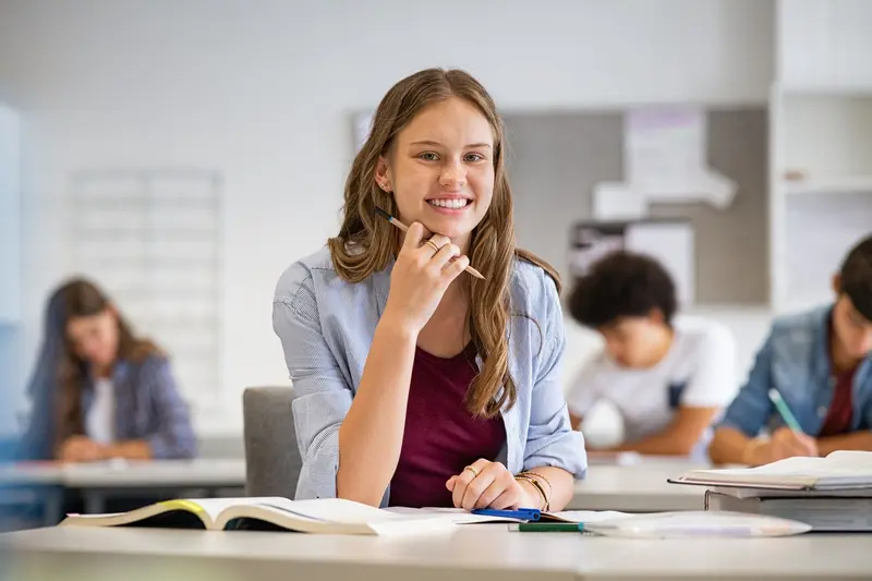 A smiling student sits at a classroom desk with an open textbook while classmates work behind her.