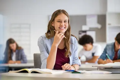 A smiling student sits at a classroom desk with an open textbook while classmates work behind her.