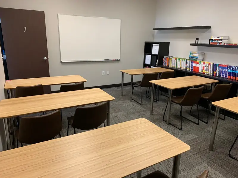 Empty classroom with wooden tables, brown chairs, a whiteboard, and shelves of colorful books along the wall.