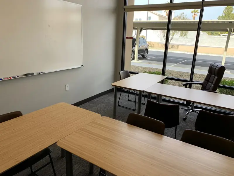 Empty classroom with wooden tables, chairs, a whiteboard, and large windows overlooking a parking lot
