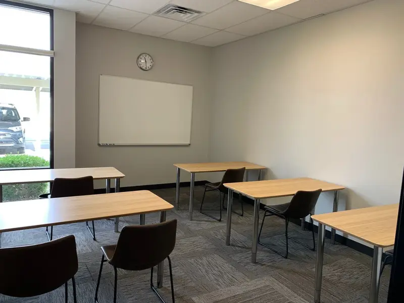 Empty classroom with wooden tables, dark chairs, a whiteboard and wall clock, and a window overlooking a parking lot.