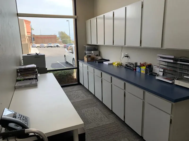 Office workroom with white cabinets above a blue countertop, a long white table, and a window overlooking a parking lot.