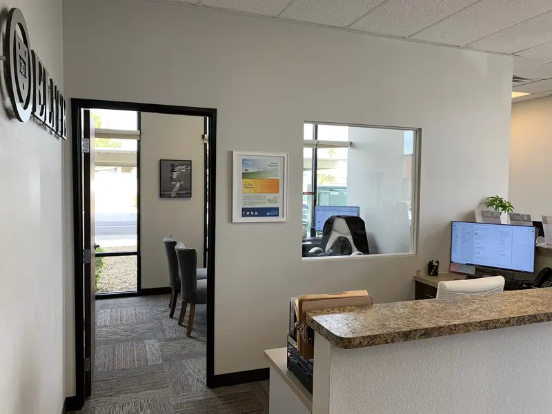 Office interior with a front desk, computer monitor, interior window and a doorway leading to a waiting area with chairs.