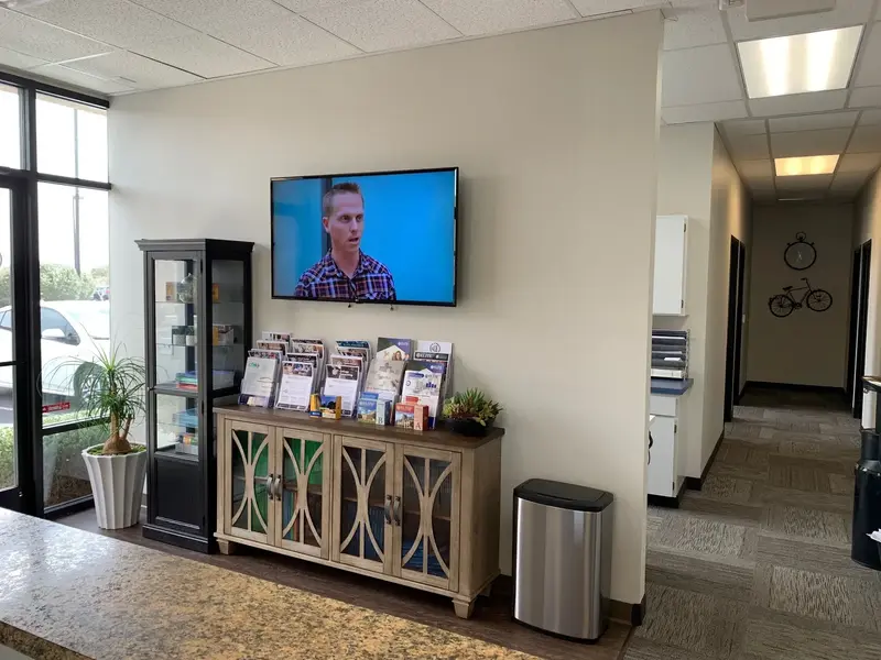 Office lobby with wall-mounted TV, brochure display on a cabinet, glass shelving unit, potted plant and hallway