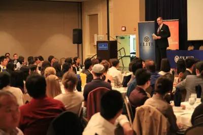 A man in a suit speaks at a podium beside an Elite banner before a large seated audience in a banquet hall.