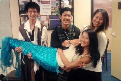 Three smiling young adults lift a laughing woman wearing a sparkly turquoise mermaid tail in an office.