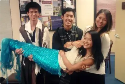 Three smiling young adults lift a laughing woman wearing a sparkly turquoise mermaid tail in an office.