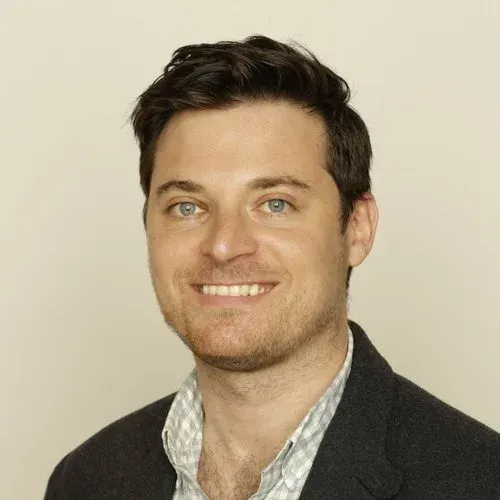 Smiling man with dark hair in a gray blazer and checked shirt against a cream background
