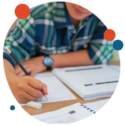A student in a plaid shirt writing with a pencil in a notebook at a desk with papers and an eraser