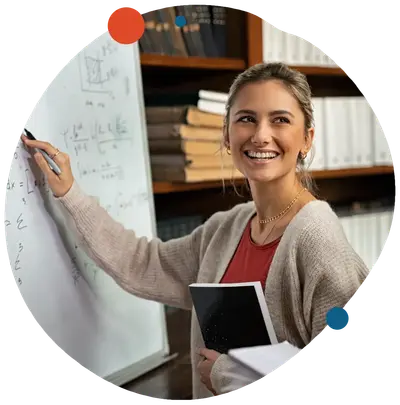 Smiling teacher writing math equations on a whiteboard while holding a notebook, bookshelves behind her