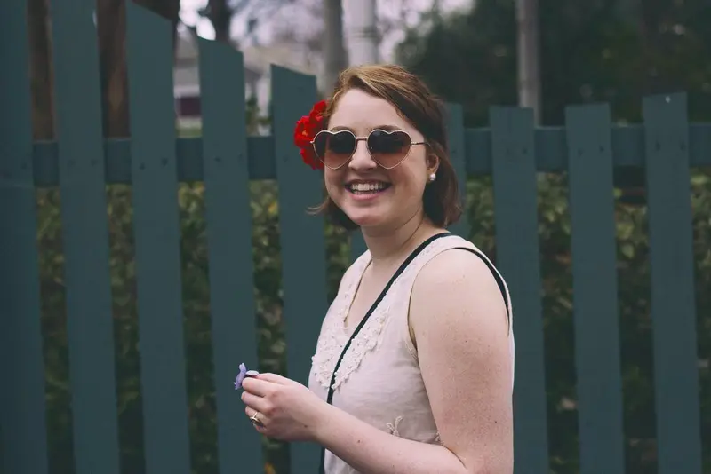 Smiling young woman in heart-shaped sunglasses with a red flower in her hair, standing by a teal picket fence