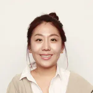 머리를 묶고 흰색 셔츠와 베이지색 가디건을 입은 채 미소 짓고 있는 여성